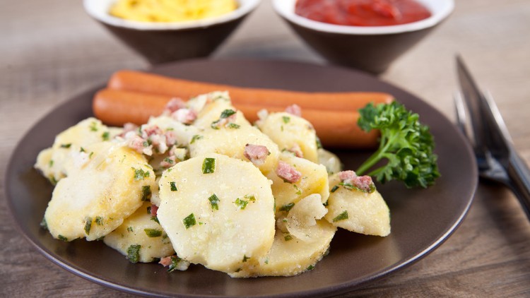 Brauner Teller mit Kartoffelsalat, zwei Würstchen und Petersilie, daneben Schalen mit gelbem Senf und rotem Ketchup.