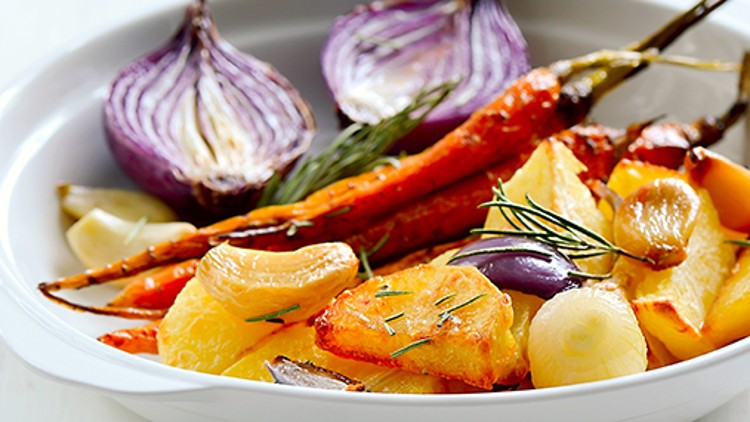 Geröstete Kartoffelscheiben, Möhren, rote Zwiebelhälften und Knoblauchzehen mit frischen Kräutern in einer weißen Schale.