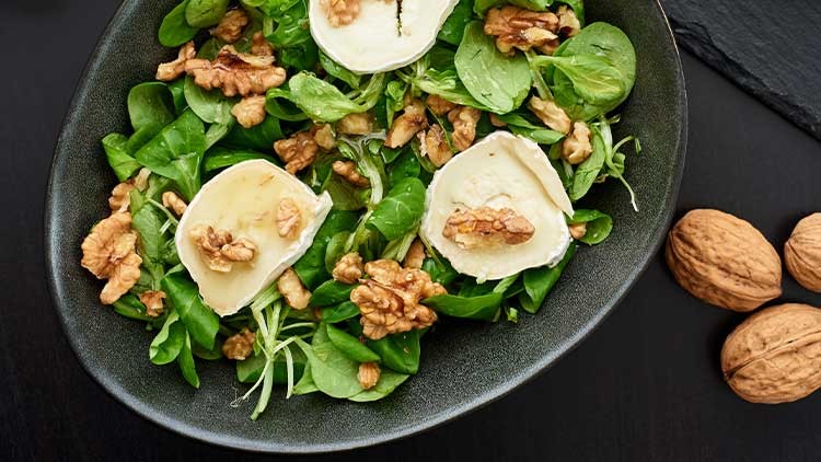 Frischer Feldsalat mit Ziegenkäsetalern und Walnüssen in einer schwarzen Schale, daneben ganze Walnüsse auf dunklem Untergrund.