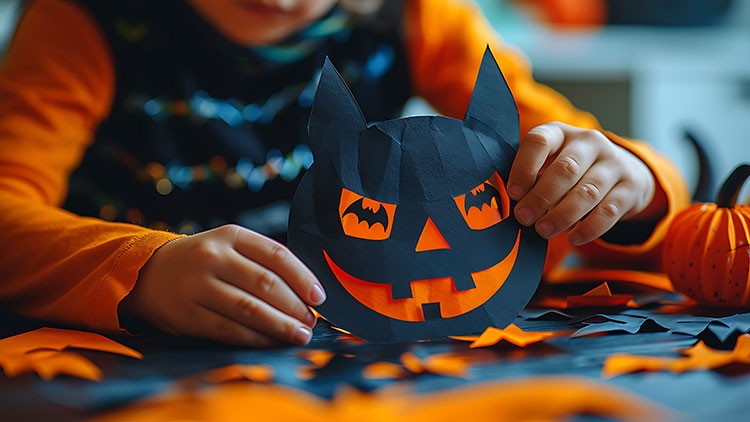 Kind hält eine schwarze Papiermaske mit orangefarbenem Kürbisgesicht, weitere Halloween-Deko liegt auf dem Tisch.
