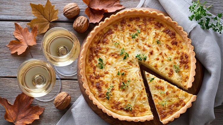 Eine goldbraune Quiche mit Kräutern liegt auf einem Holztisch, daneben zwei Gläser Weißwein, Walnüsse, Herbstblätter und ein graues Tuch.