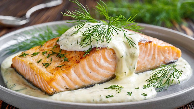 Gegrilltes Lachsfilet mit heller Dill-Sauce und frischen Kräutern auf einem grauen Teller, im Hintergrund grüne Dekoration.