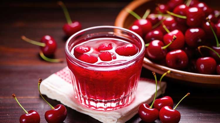 Ein Glas rot leuchtender Kirschsaft mit Kirschen darin steht auf einem Stofftuch, daneben liegen frische Kirschen und eine Holzschale.