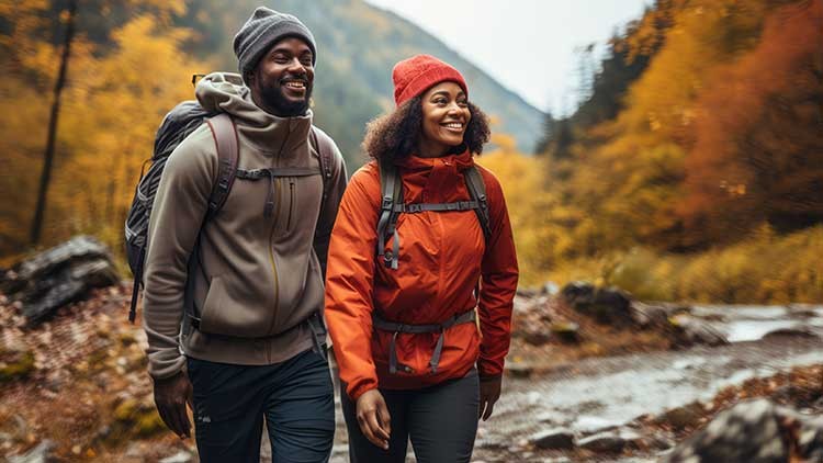 Zwei Personen in Outdoor-Kleidung mit Rucksäcken wandern lächelnd auf einem Waldweg im Herbst.