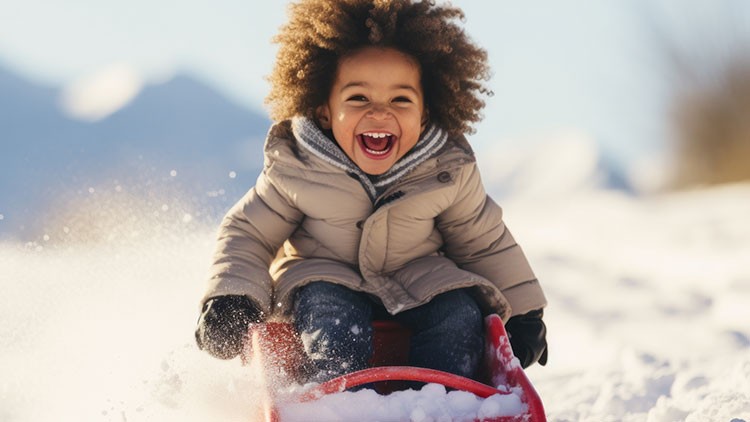 Kind in heller Winterjacke und Handschuhen fährt lachend auf einem roten Schlitten durch schneebedeckte Landschaft.