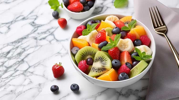 Bunte Obstsalatschale mit Kiwi, Erdbeeren, Blaubeeren, Banane und Minzblättern auf Marmortisch neben goldener Gabel.