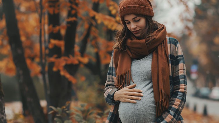 Eine Person in Herbstkleidung steht im Wald, hält die Hände auf den Bauch, umgeben von orangefarbenen Blättern.