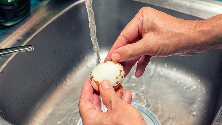 Zwei Hände schälen ein gekochtes Ei unter fließendem Wasser über einem Edelstahl-Spülbecken.