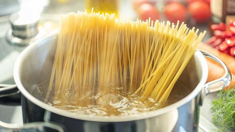 Lange, ungekochte Spaghetti stehen in einem Kochtopf mit kochendem Wasser, im Hintergrund sind Tomaten und Kräuter.