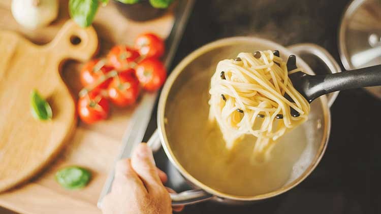 Eine Hand hebt gekochte Spaghetti mit einem schwarzen Schaumlöffel aus einem Topf, im Hintergrund Tomaten und Basilikum.