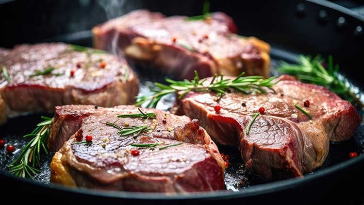 Vier Stücke Fleisch mit Kräutern und roten Pfefferkörnern braten in einer schwarzen Pfanne.