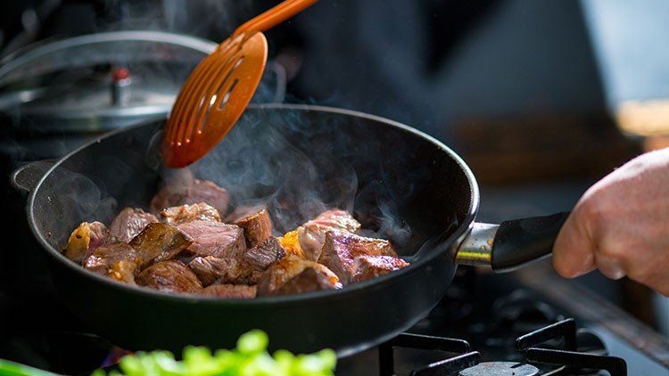 In einer Pfanne werden auf einem Herdstück angebraten, während ein orangefarbener Pfannenwender Fleischstücke bewegt .
