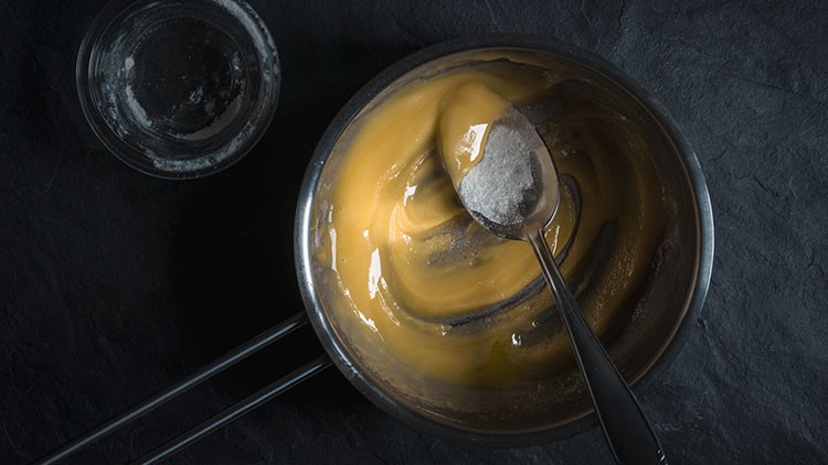 Ein Löffel mit weißem Pulver liegt in einem kleinen Topf mit gelblicher Creme, daneben steht ein Glas mit Pulverresten.
