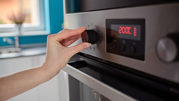 Eine Hand dreht an einem schwarzen Drehknopf eines silbernen Backofens, dessen Display „200°C“ anzeigt.