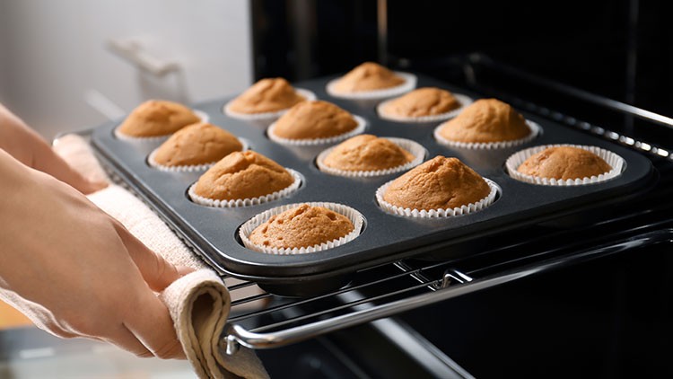 Ein Backblech mit zwölf goldbraunen Muffins in Papierförmchen wird aus einem Ofen herausgezogen.
