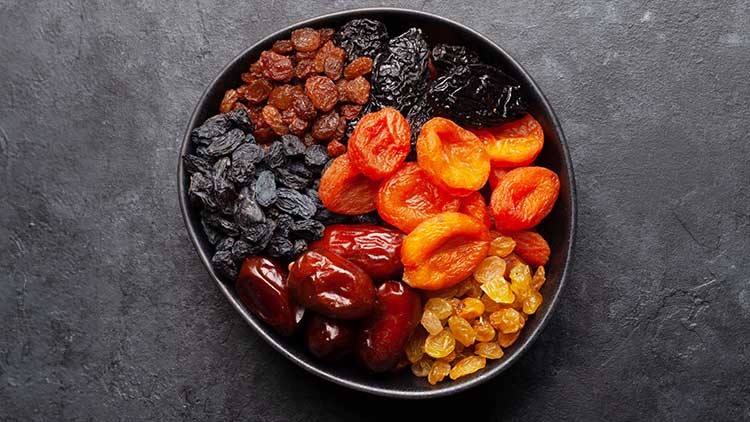 Schwarze Schale mit getrockneten Früchten wie Aprikosen, Datteln, Rosinen, Pflaumen und dunklen Beeren auf grauem Untergrund.