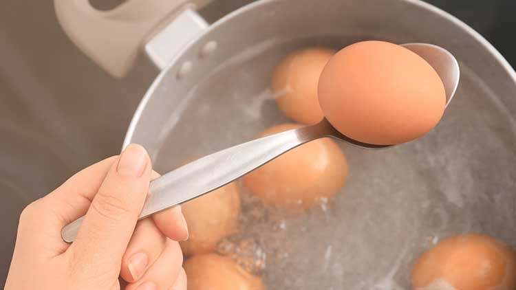 Eine Hand hält einen Löffel mit einem braunen Ei über einem Topf mit kochendem Wasser und weiteren Eiern.