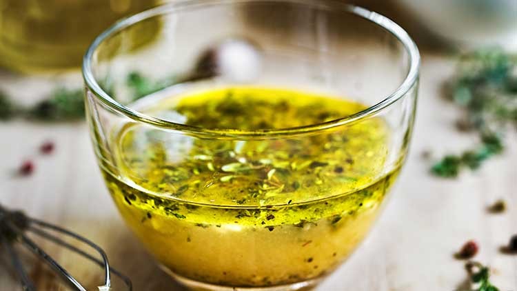 Kleiner Glasbehälter mit Kräuterdressing aus Öl und Kräutern, im Hintergrund unscharfe Gewürze und Küchenutensilien.