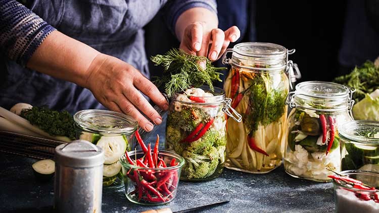 Eine Person füllt Einmachgläser mit Gemüse, Kräutern und Gewürzen wie Chili, daneben liegen weitere Zutaten auf dem Tisch.