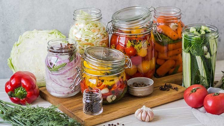 Verschiedene Einmachgläser mit eingelegtem Gemüse wie Paprika, Karotten und Gurken stehen auf einem Holzbrett mit Kräutern.