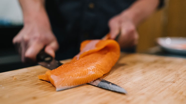 Eine Person filetiert ein Stück frischen Lachs mit einem Messer auf einem Holzbrett.