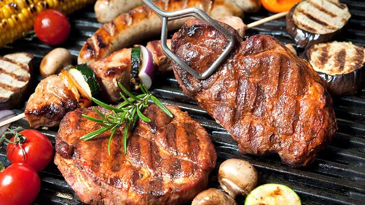 Gegrilltes Fleisch, Würstchen, Gemüse wie Zucchini und Paprika sowie Maiskolben und Kirschtomaten auf einem Grill.