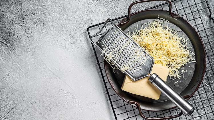 Eine Metallreibe liegt auf einer schwarzen Pfanne, daneben ein Stück Käse und frisch geriebene Käsestreifen.