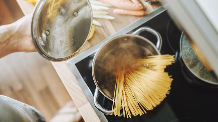Ein Kochtopf mit kochendem Wasser und ungekochten Spaghetti steht auf einem Herd, daneben liegen Karotten und Spargel.