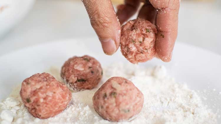 Eine Hand rollt aus gewürztem Hackfleisch eine kleine Kugel und wendet sie in Mehl auf einem weißen Teller.
