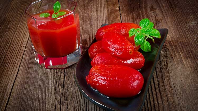 Ein Glas Tomatensaft mit Basilikum und ein schwarzer Teller mit geschälten Tomaten und Basilikumblättern auf Holztisch.