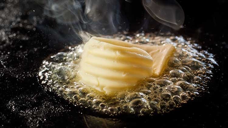 Ein Stück Butter schmilzt zischend in heißem Fett auf einer dunklen Bratfläche, Dampf steigt auf.