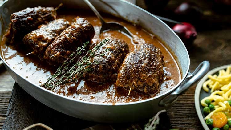 Vier Rinderrouladen in brauner Soße mit Thymianzweigen in einer ovalen Metallschale, daneben ein Teller mit Spätzle und Gemüse.