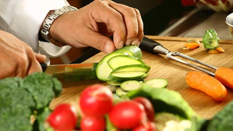 Hände schneiden mit einem Messer grüne Zucchini auf einem Holzbrett, daneben liegen Brokkoli, Radieschen und eine Karotte.