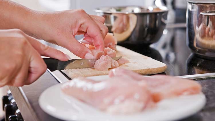 Hände schneiden rohes Hähnchenfleisch auf einem Holzbrett in Würfel, im Hintergrund stehen Schüsseln und Töpfe.