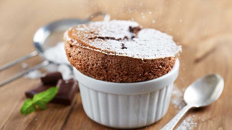 Ein Schokoladen-Soufflé in einer weißen Keramikform, bestäubt mit Puderzucker, daneben ein Löffel und Schokoladenstücke.