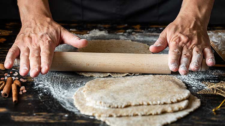 Hände rollen mit einem Holz-Nudelholz Teig auf einer bemehlten Oberfläche aus, umgeben von Mehl, Zimtstangen und Salz.