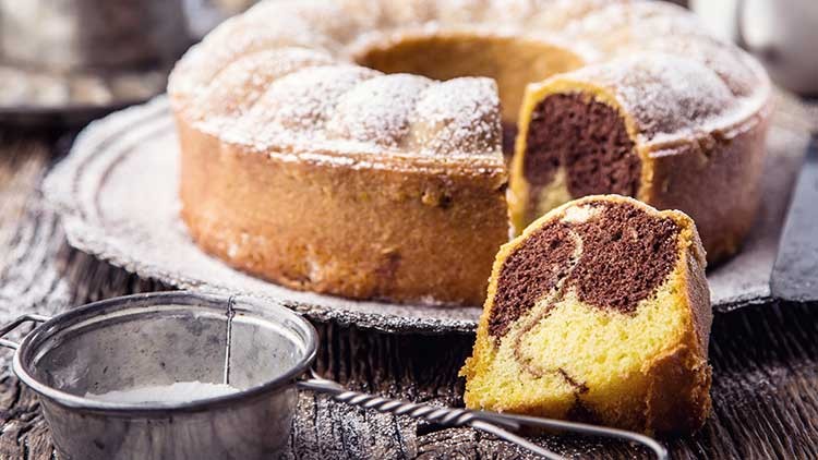 Ein Marmorkuchen mit Puderzucker liegt auf einem Teller, daneben ein angeschnittenes Stück und ein Sieb mit Puderzucker.