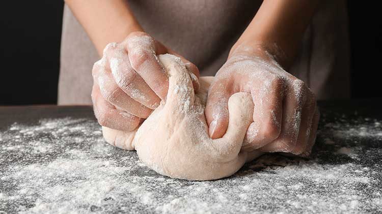 Hände kneten einen bemehlten Teig auf einer mit Mehl bestreuten dunklen Arbeitsfläche.