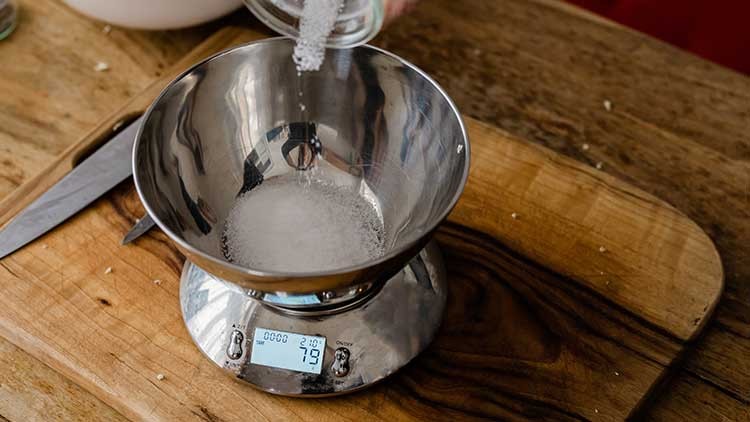 Metallene Küchenwaage mit digitaler Anzeige wiegt Zucker in einer Edelstahlschüssel auf einem Holzbrett ab.
