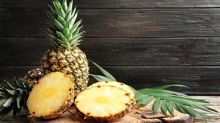 Eine ganze Ananas steht auf einem Holzbrett, daneben zwei halbierte Ananasscheiben und grüne Blätter, Hintergrund aus dunklem Holz.