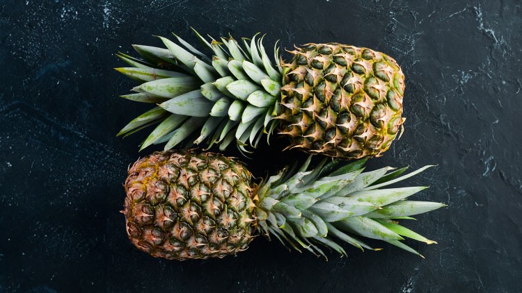 Zwei ganze Ananas mit grünem Strunk liegen nebeneinander auf schwarzem Untergrund.