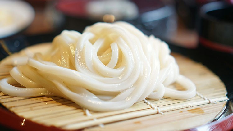 Gekochte, weiße Udon-Nudeln liegen aufgerollt auf einer Bambusmatte mit unscharfem Hintergrund.