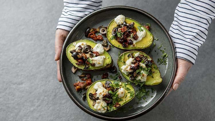 Vier Avocadohälften mit Käsecreme, schwarzen Oliven, getrockneten Tomaten und Kräutern auf einem Teller, gehalten von zwei Händen.