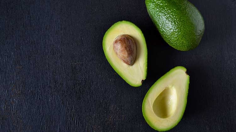 Eine ganze Avocado und zwei Hälften, eine mit Kern, liegen auf einer dunklen, strukturierten Oberfläche.