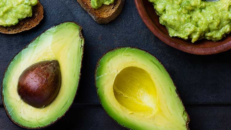 Halbierte Avocado mit Kern, Avocadohälfte ohne Kern, Avocadocreme in Schale und Avocadocreme auf Brot auf dunklem Untergrund.