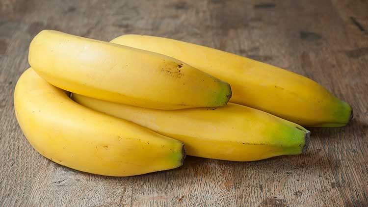 Vier gelbe Bananen liegen auf einer Holzoberfläche, teils mit grünen Spitzen.