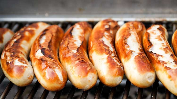 Sechs hellbraun gegrillte Würstchen mit Röststreifen liegen nebeneinander auf einem silbernen Grillrost.