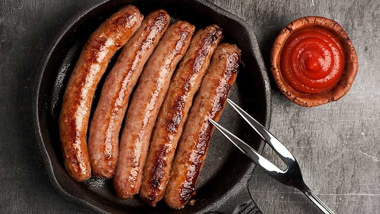 Fünf gebratene Bratwürste liegen in einer gusseisernen Pfanne, daneben eine kleine Schale mit rotem Ketchup und eine Fleischgabel.