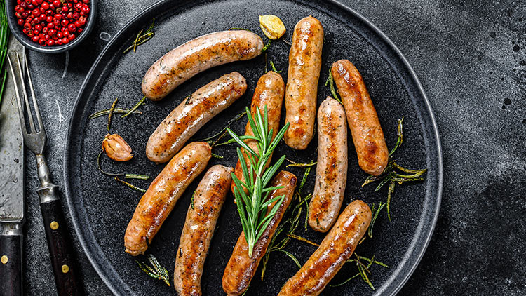 Mehrere gebratene Würstchen und Zweige frischen Rosmarins auf einem dunklen Teller mit Knoblauch und Besteck daneben.