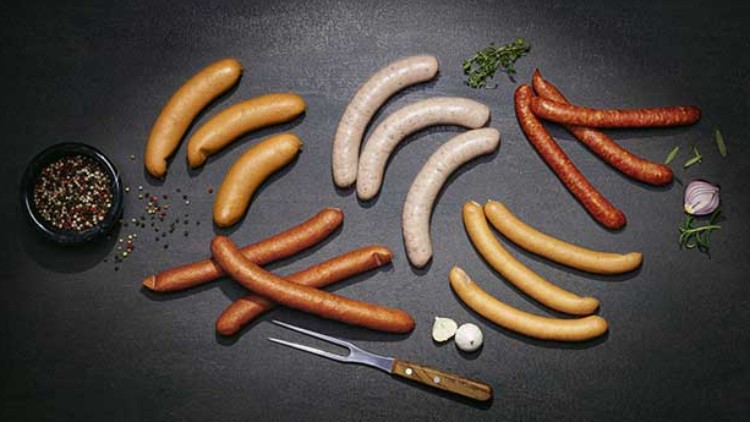 Verschiedene Würstchen liegen auf einer dunklen Oberfläche mit Gewürzen, frischen Kräutern, Knoblauch und einer Fleischgabel.
